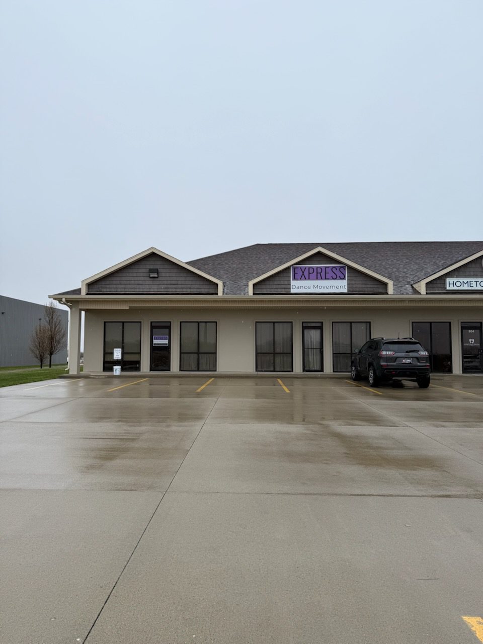 Express Dance Movement storefront with a gray sky backdrop. The building features a prominent purple sign, and the parking lot appears wet.
