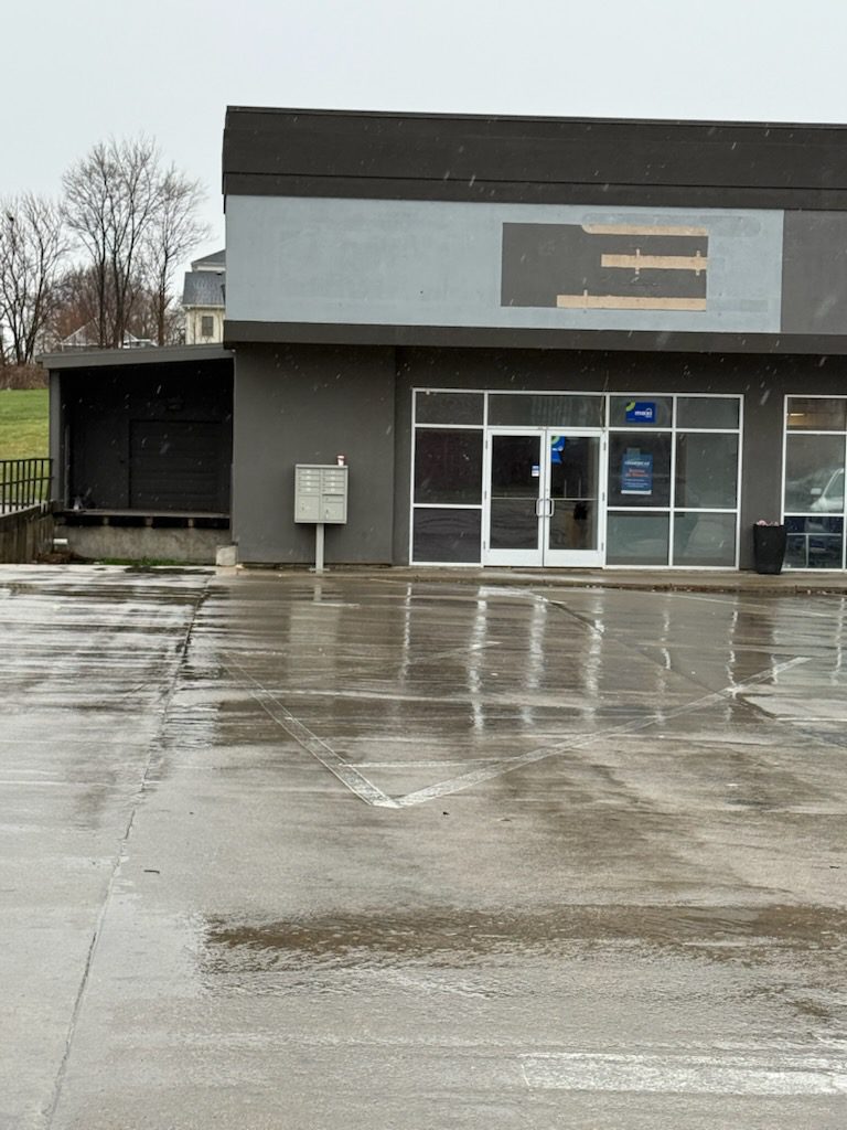Empty commercial space with wet pavement, featuring a glass entrance and a mailbox. Construction work is indicated by covered signage.